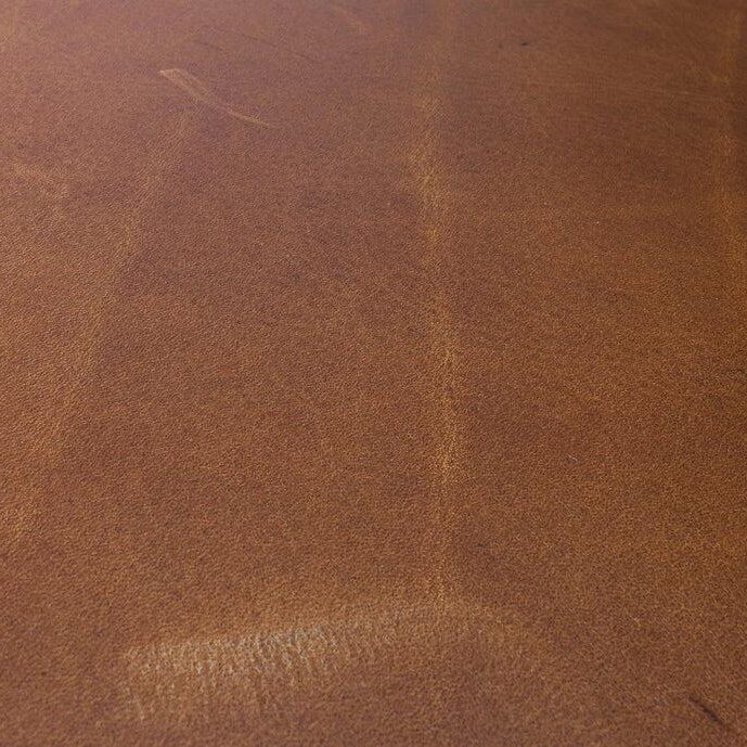 Close-up of a medium brown full-grain leather surface showing subtle natural grain lines, tonal shifts, and a faint scratch mark in the upper portion